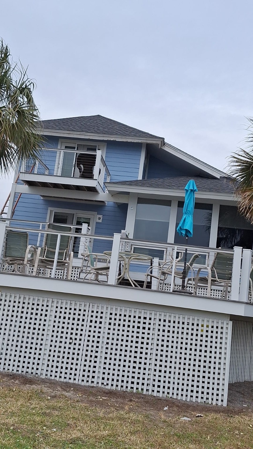 Rear of the blue coastal house with white trim, upper balcony and deck railings